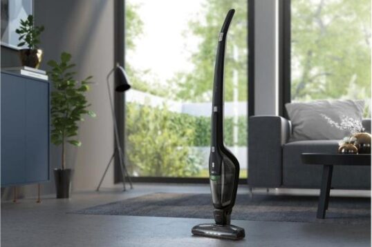 Een moderne staande stofzuiger staat in een stijlvolle woonkamer en is klaar voor gebruik. Achtergrond met veel licht, planten en een comfortabele bank dragen bij aan de huiselijke sfeer.
