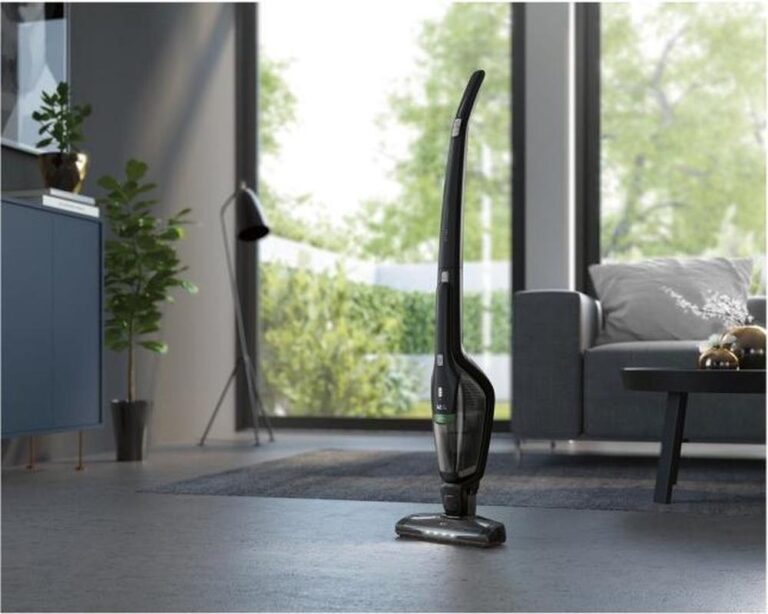 Een moderne staande stofzuiger staat in een stijlvolle woonkamer en is klaar voor gebruik. Achtergrond met veel licht, planten en een comfortabele bank dragen bij aan de huiselijke sfeer.
