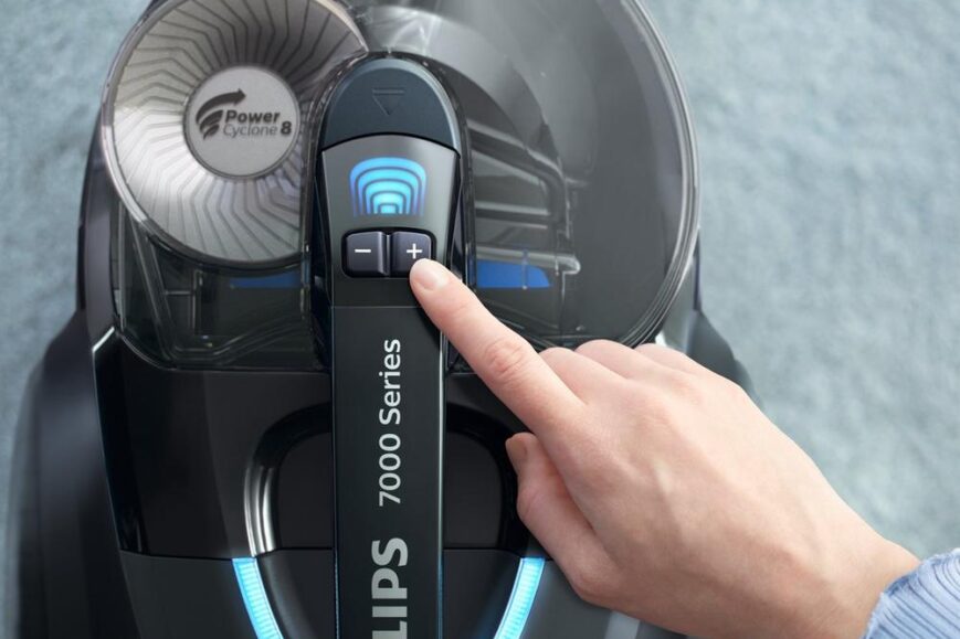 Een close-up van een hand die een knop op een Philips 7000 Series stofzuiger bedient. De afbeelding toont de bedieningspanelen en het ontworpen stofreservoir van het apparaat.