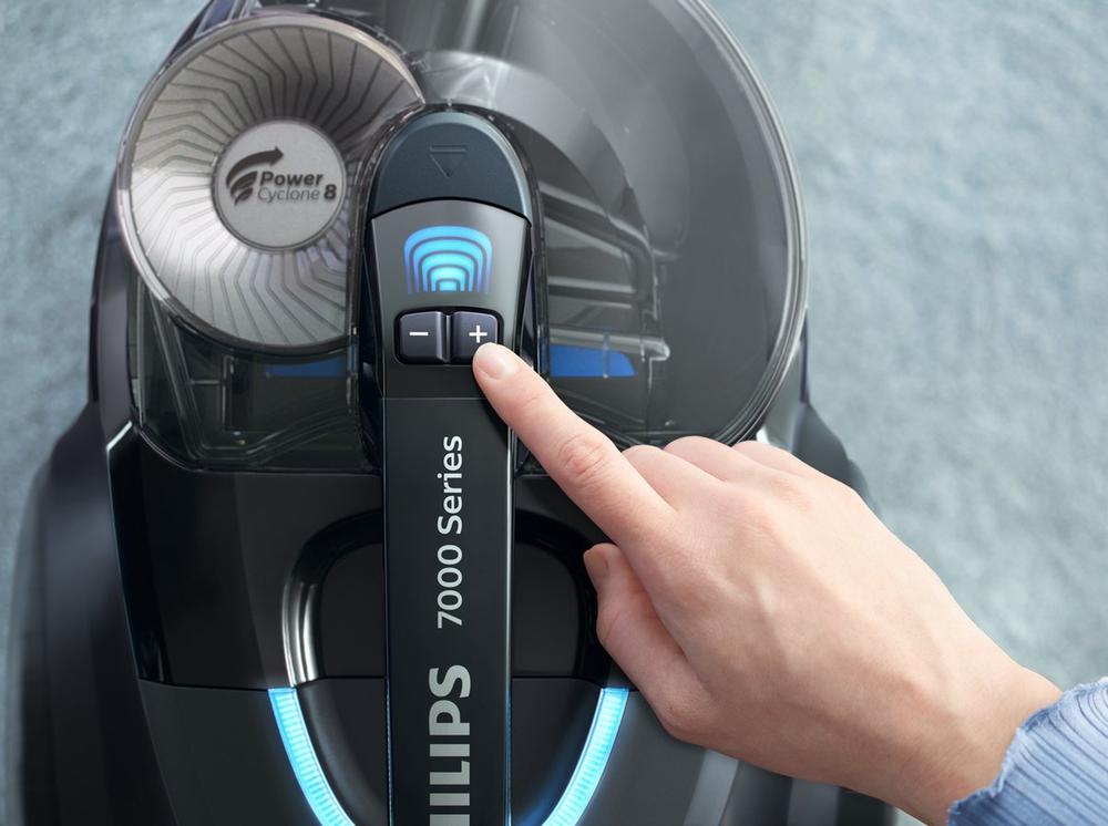 Een close-up van een hand die een knop op een Philips 7000 Series stofzuiger bedient. De afbeelding toont de bedieningspanelen en het ontworpen stofreservoir van het apparaat.