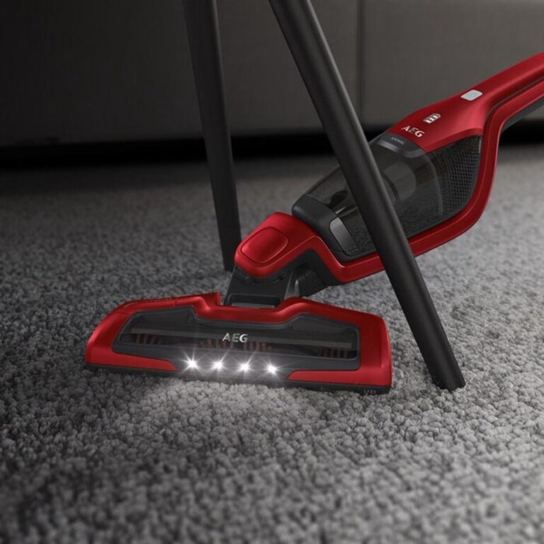 Een rode stofzuiger van AEG met een verlicht mondstuk, geplaatst onder een meubelstuk. De stofzuiger staat op een grijze, zachte vloerbedekking, wat duidt op gebruik voor het schoonmaken van tapijt.