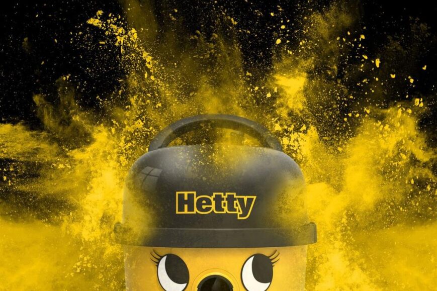 Afbeelding van een vrolijke gele stofzuiger met een zwart bovenstuk, genaamd "Hetty". De achtergrond bevat een explosie van gele kleurstof, die een dynamisch en levendig effect creëert.