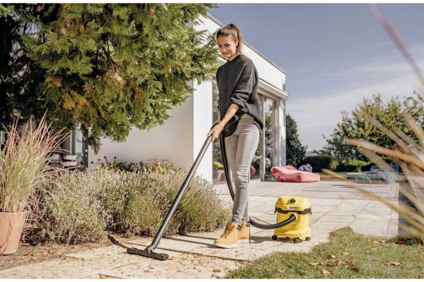 Een vrouw met een grijze trui en beige laarzen gebruikt een stofzuiger op een terras, omringd door planten en bomen. Op de achtergrond zijn een modern huis en kleurrijke meubels zichtbaar.