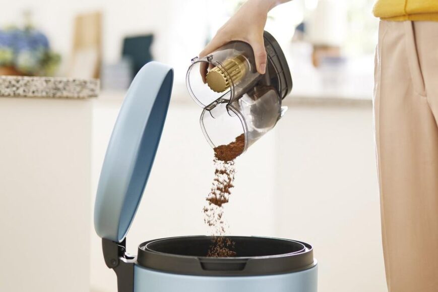 Een hand giet gemalen koffie in een blauwe prullenbak. De afbeelding illustreert het afvalproces na het zetten van koffie in een moderne keukenomgeving.