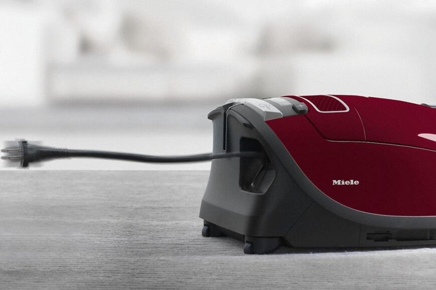 Een rode Miele stofzuiger staat op een tapijt, met de stekker zichtbaar die uit de behuizing komt. De achtergrond is vaag, wat de focus op de stofzuiger legt.