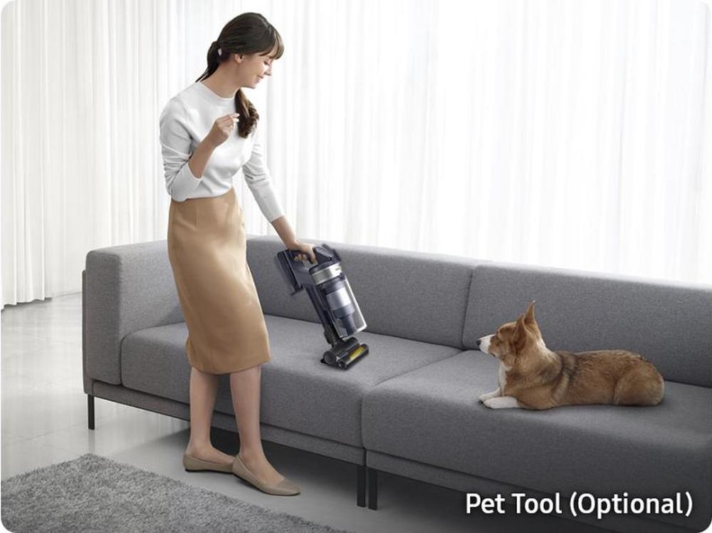 Een vrouw in een witte trui en een beige rok stofzuigt een grijze bank met een handstofzuiger terwijl een hond op de bank sitzt. De afbeelding illustreert het gebruik van een optionele pet tool die speciaal is ontworpen voor het verwijderen van dierenharen.