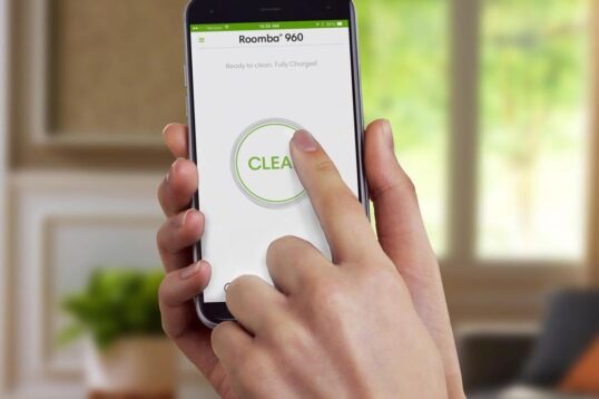 Een hand raakt het scherm van een smartphone met de Roomba 960-app, waarbij de "CLEAR" knop wordt ingedrukt. De achtergrond toont een moderne woonkamer met veel natuurlijk licht en planten.
