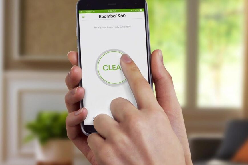 Een hand raakt het scherm van een smartphone met de Roomba 960-app, waarbij de "CLEAR" knop wordt ingedrukt. De achtergrond toont een moderne woonkamer met veel natuurlijk licht en planten.