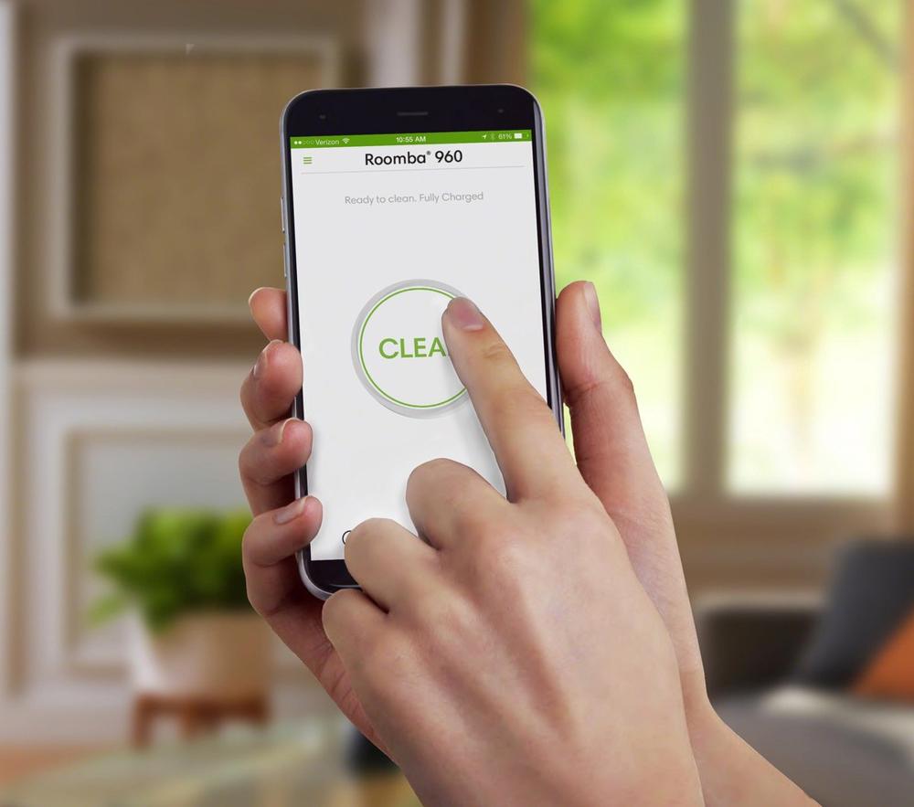Een hand raakt het scherm van een smartphone met de Roomba 960-app, waarbij de "CLEAR" knop wordt ingedrukt. De achtergrond toont een moderne woonkamer met veel natuurlijk licht en planten.