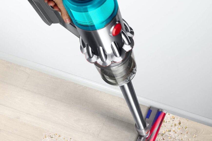 Een handstofzuiger wordt gebruikt om korrels en stof van een houten vloer op te zuigen. De stofzuiger heeft een moderne uitstraling met een helderblauwe bovenkant en een transparante opvangbak.