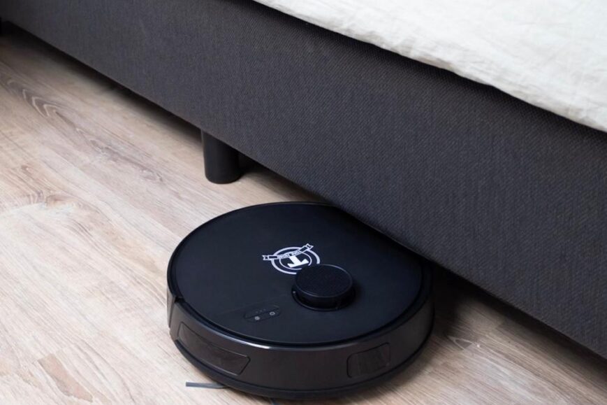 Een zwarte robotstofzuiger staat onder een bed en reinigt de vloer. De afbeelding benadrukt de effectiviteit van het apparaat in moeilijk bereikbare hoeken en onder meubelen.