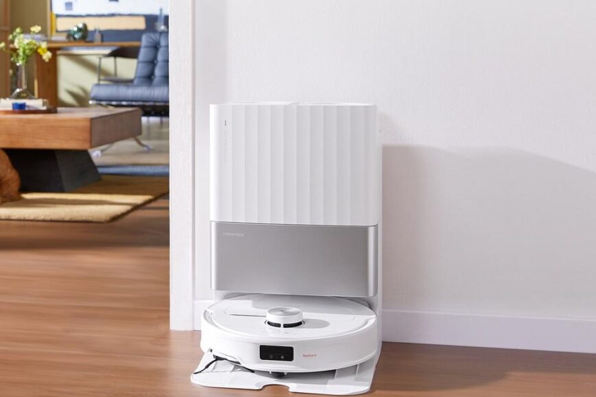 Afbeelding van de Roborock Qrevo Master robotstofzuiger, geplaatst op een houten vloer naast een wand. De stofzuiger staat voor een moderne woonkamer met meubilair op de achtergrond.