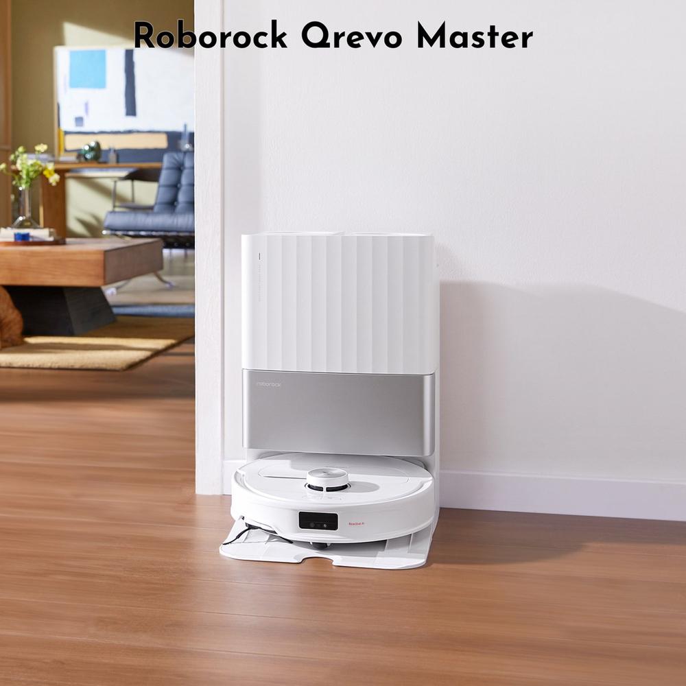 Afbeelding van de Roborock Qrevo Master robotstofzuiger, geplaatst op een houten vloer naast een wand. De stofzuiger staat voor een moderne woonkamer met meubilair op de achtergrond.