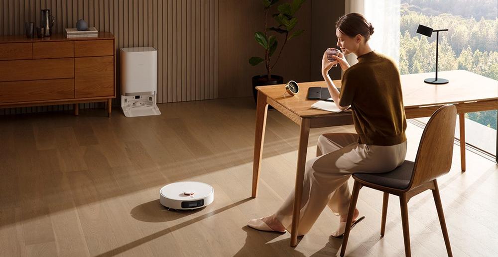 Een vrouw zit aan een houten tafel in een moderne kamer en drinkt een kopje. Een automatische stofzuiger rolt over de vloer in de buurt van de tafel, terwijl er een grote raam met uitzicht op een bosachtige omgeving zichtbaar is.
