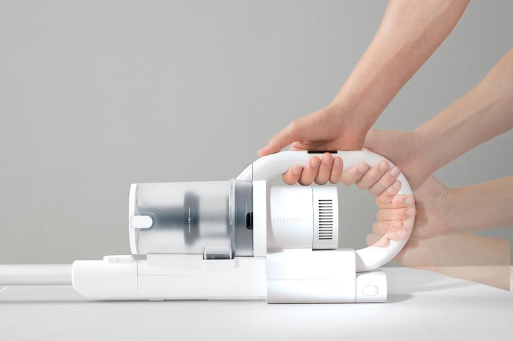 Afbeelding van een draadloze stofzuiger met een transparante stofreservoir en een witte behuizing, waarbij twee handen de stofzuiger vasthouden. De nadruk ligt op de ergonomische grip en het moderne ontwerp, wat de functionaliteit van het apparaat benadrukt.