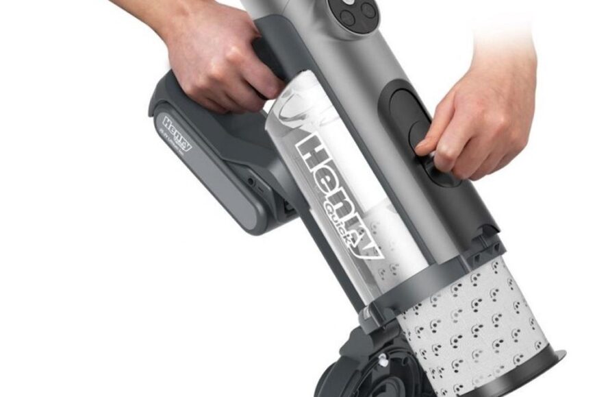 Draagbare Henry stofzuiger in grijstinten, vastgehouden door een hand die de aan-/uitknop bedient. De transparante stofcontainer toont het filter en de opbouw van de stofzuiger.
