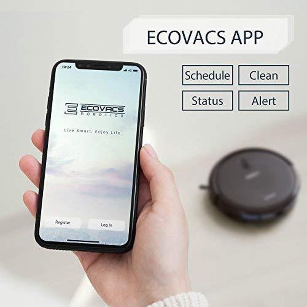 Een hand houdt een smartphone vast die de ECOVACS-app toont, met opties voor registreren en inloggen. Op de achtergrond is een robotstofzuiger zichtbaar, wat duidt op het gebruik van de app voor het beheren van schoonmaaktaken.