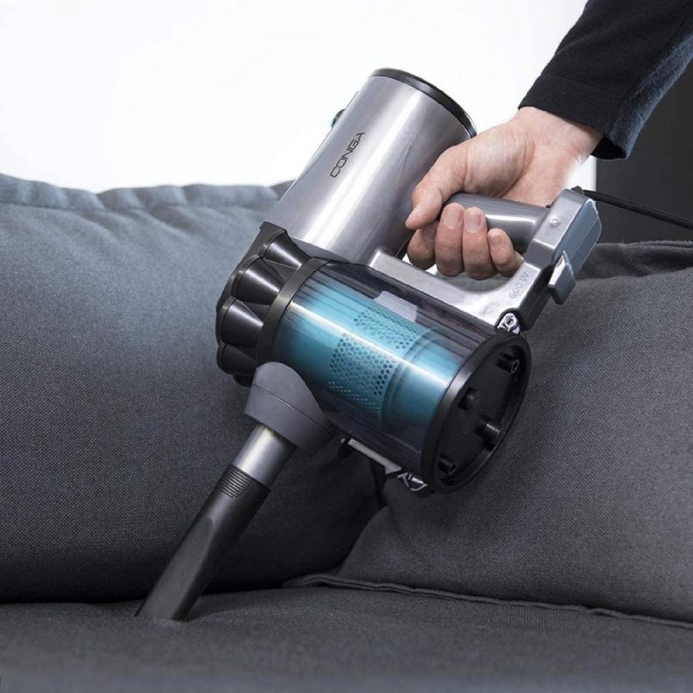 Een persoon gebruikt een handstofzuiger om een bank schoon te maken. De stofzuiger heeft een zilverkleurig ontwerp met een transparante opvangbak.