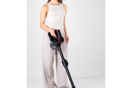 Een vrouw in een casual outfit houdt een draadloze stofzuiger vast en glimlacht terwijl ze staat op een witte vloer. De stofzuiger heeft een modern ontwerp met een zwarte en rode kleurstelling, wat de focus legt op gebruiksgemak en functionaliteit.