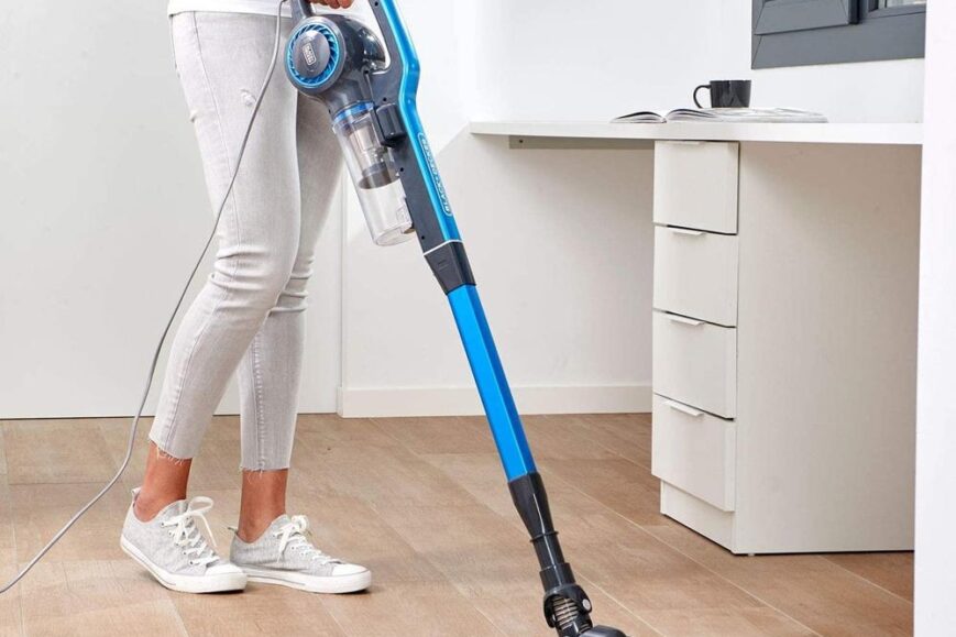 Een vrouw gebruikt een blauwe, draadloze stofzuiger terwijl ze op een houten vloer staat. Op de achtergrond is een werkruimte zichtbaar met een bureau en een kopje koffie.