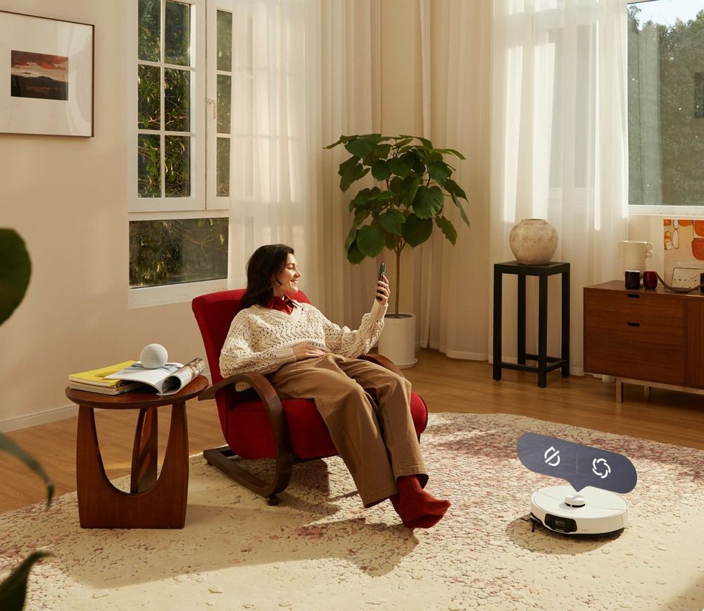 Een vrouw zit ontspannen in een rode schommelstoel en kijkt naar haar smartphone, terwijl een robotstofzuiger de vloer schoonmaakt. Op de achtergrond zijn een plant, een bijzettafel en grote ramen zichtbaar die natuurlijk licht binnenlaten.